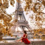 Kulla Eiffel në Paris mes pemëve me gjethe të arta vjeshte, me një grua me fustan të kuq në plan të parë.