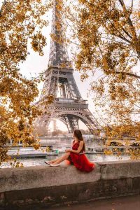 Kulla Eiffel në Paris mes pemëve me gjethe të arta vjeshte, me një grua me fustan të kuq në plan të parë.
