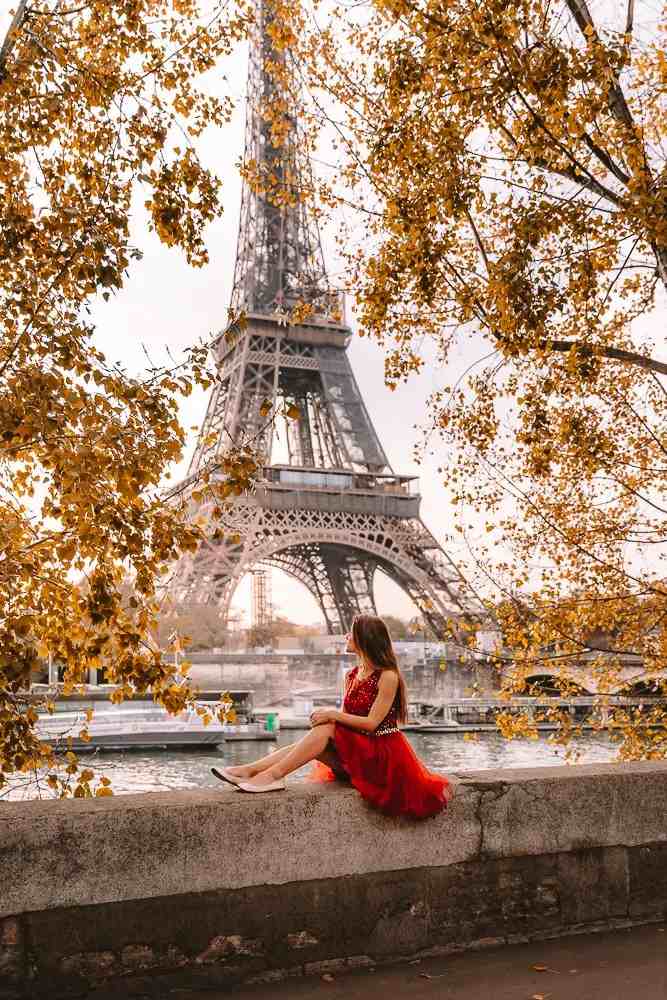 Kulla Eiffel në Paris mes pemëve me gjethe të arta vjeshte, me një grua me fustan të kuq në plan të parë.